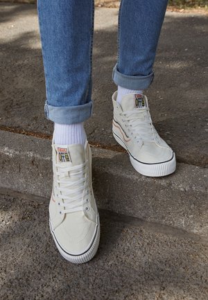 Nogi w podwiniętych niebieskich dżinsach i białych wysokich trampkach Vans z białymi skarpetami, stojące na szarym asfalcie i krawężniku.