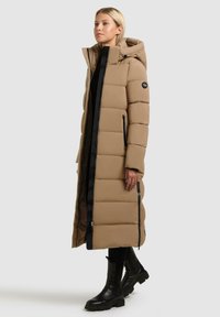 Lange, beige pufferjas met een capuchon, met horizontale stiknaden, zwarte ritssluitingen en zijzakken. Gecombineerd met zwarte enkellaarsjes.