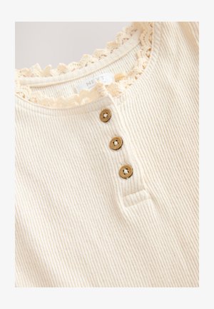 Šviesiai smėlio spalvos briaunotas viršus su nėrinių apdaila, priekyje trys medinės sagos. Minkšta tekstūra ir kasdieniškas dizainas.