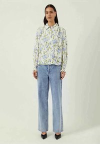 Camisa floral de botão em branco com flores azuis e verdes, combinada com jeans de corte reto em lavagem clara e sapatos de salto preto com bico fino.