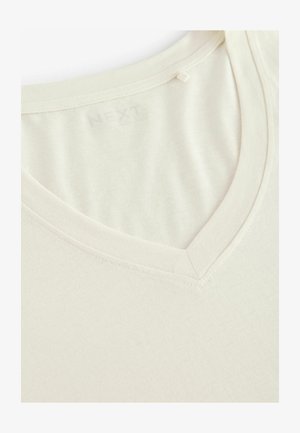 Camiseta de cuello en V crema hecha de tela suave. Presenta una textura lisa, un diseño sencillo y bordes cosidos con una etiqueta de marca sutil en el cuello.