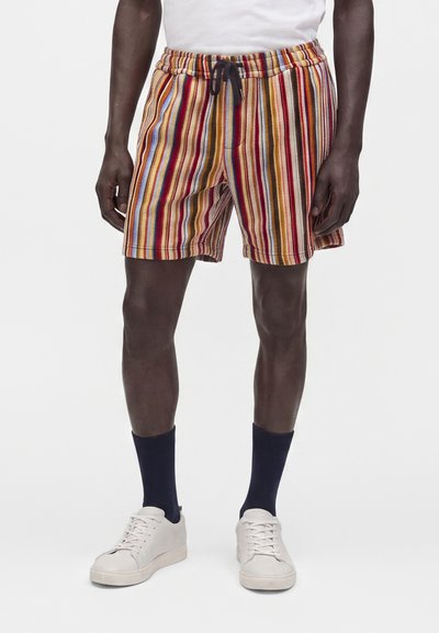 Homme portant un short à rayures verticales multicolores avec un cordon noir, des chaussettes bleu marine, des baskets blanches et une chemise blanche.