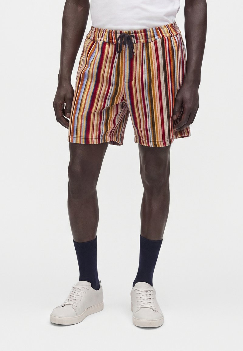 Homme portant un short à rayures verticales multicolores avec un cordon noir, des chaussettes bleu marine, des baskets blanches et une chemise blanche.
