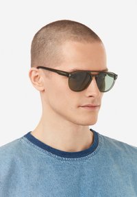 Aurinkolasit, joissa on tummanvihreät linssit ja musta metallinen kehys. Minimalistinen muotoilu, jossa on kaksoissilta. Malli pukeutuneena farkkupaitaan.