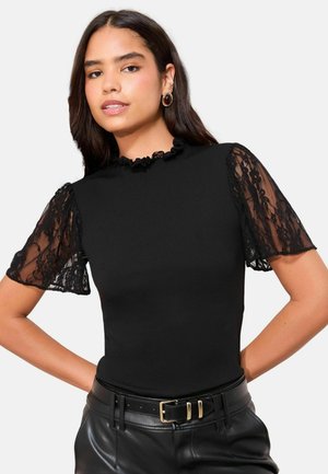 Schwarzes tailliertes Oberteil mit kurzen Spitzenärmeln und einem rüschierten Ausschnitt, kombiniert mit hoch taillierten Lederhosen, die aufwendige Spitzenakzente zeigen.