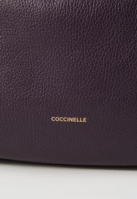 Borsa in pelle viola scuro con superficie testurizzata. Logo "COCCINELLE" in oro a caldo nell'angolo in basso a destra.