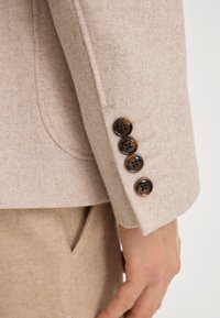 Rękaw beżowej wełnianej marynarki z czterema ciemnymi okrągłymi guzikami. Tkanina ma teksturowany wygląd z widocznym przeszyciem i klasycznym wcięciem na klapie.