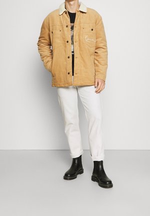 Hombre con chaqueta de pana beige con cuello de borrego blanco, camiseta gráfica negra, pantalones blancos y botas de cuero negras, de pie contra una pared gris.