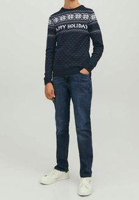 Námořnický svetr s bílým vánočním vzorem a nápisem "ŠŤASTNÉ VÁNOCE", pořízený k tmavým džínům a bílým teniskám se zelenými akcenty.