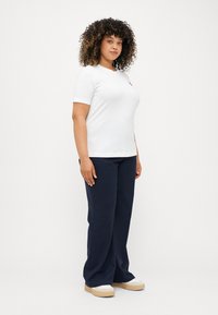 Λευκό κοντομάνικο μπλουζάκι εφαρμοστό, σε συνδυασμό με ναυτικό μπλε παντελόνι φαρδύ και μπεζ αθλητικά παπούτσια με λευκές λεπτομέρειες. Γενικά λεία υφή.