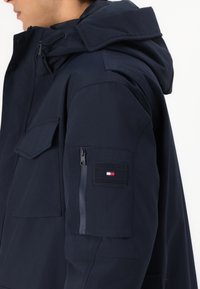 Veste bleu marine avec un col montant, une capuche, de multiples poches et une fermeture éclair. Dispose d'un patch sur le bras gauche avec le logo et des accents de couleur.