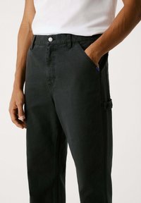 Schwarze Baumwoll-Cargo-Hosen mit lässiger Passform, ausgestattet mit vorderen Taschen und einer Seitentasche. Sichtbare Nähte und ein KnopfverschlussDETAIL.