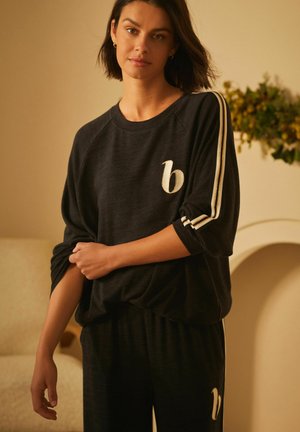 Zwart loungewear-set met een relaxte top die een witte geborduurde 'b' logo en witte zijstreepjes heeft. Zachte textuur, casual ontwerp.