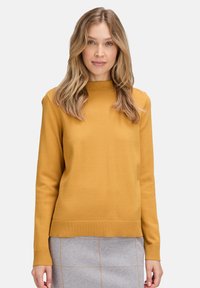 Mustár sárga, hosszú ujjú pulóver bordázott nyakkivágással és mandzsettákkal, sima tapintású és laza fazonú, szürke mintás szoknyával párosítva.