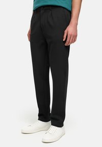 Pantaloni neri su misura con una vestibilità aderente, realizzati in un tessuto liscio, con pieghe frontali e tasche laterali, abbinati a sneaker bianche.