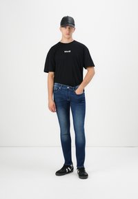 Joven con una gorra negra, camiseta negra con texto blanco, jeans ajustados azules y zapatillas negras, de pie contra un fondo blanco.