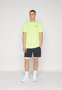 Neon žluté sportovní tričko s krátkým rukávem a texturovanou tkaninou, spárované s tmavými šortkami s bílou podšívkou a logem. Bílé boty.