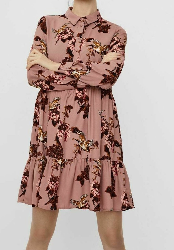 Femme portant une robe rose poussiéreuse à manches longues avec des imprimés d'oiseaux et de fleurs, debout, les bras croisés, sur un fond uni.