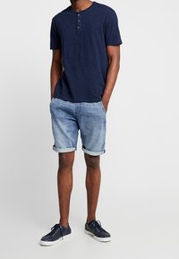 Camiseta henley de manga corta azul marino con tres botones; pantalones cortos de mezclilla azul claro con dobladillos enrollados; zapatillas deportivas azules marinas con suelas blancas.
