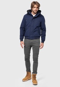 Granatowa kurtka bomberka z kapturem, zapinana na zamek błyskawiczny, z bocznymi kieszeniami, w połączeniu z szarymi spodniami slim-fit i brązowymi butami typu lace-up.