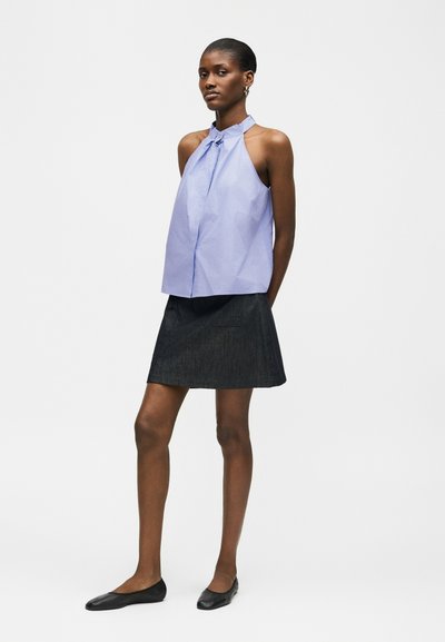 Femme debout portant un haut à col halter sans manches bleu clair associé à une jupe noire jusqu'aux genoux et des chaussures plates noires sur un fond uni.