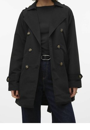 Woman wearing a black double-breasted trench coat over a black top and dark blue jeans with a black belt, standing against a white background.