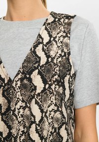 Tan en zwart slangenprint vest over een lichtgrijze T-shirt met korte mouwen. Het vest heeft een diepe V-hals en een ribbelstructuur.
