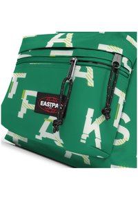 Bolsa de tela verde con un estampado blanco, cremalleras con tiradores negros y un prominente parche del logo "EASTPAK". El diseño incluye patrones abstractos.
