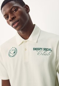 Cremefarbener Poloshirt mit Kragen, versehen mit grünen Grafiken: einem Snoopy-Design und dem Text "SNOOPY SOCIAL Club". Mit Zweiknopf-Leiste.