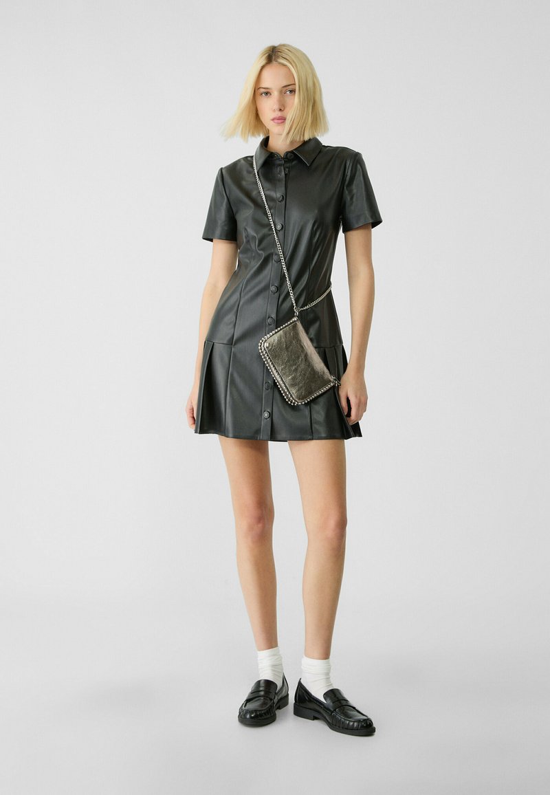 Abito in pelle nera a camicia con maniche corte, chiusura con bottoni, gonna plissettata e borsa a tracolla in catena argentata. Calzini bianchi e mocassini neri.