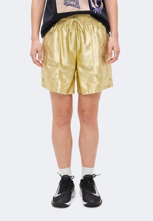 Person trägt glänzende goldene Shorts mit Kordelzug und dezentem Muster, kombiniert mit schwarzen Sneakers und weißen Socken, auf einfarbigem Hintergrund stehend.