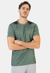 Camiseta atlética verde con mangas cortas, cuello redondo, acentos negros en los hombros y un logo en la parte frontal. Textura de tela suave.
