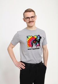 Šedé bavlněné tričko s barevným tie-dye logem MTV, které má efekt kapající barvy; nošeno s černými kalhotami a černým opaskem.