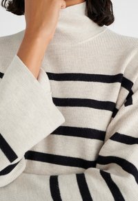 Světlý béžový svetr s námořnickými pruhy, s hřejivým rolákem a oversized rukávy, předvádějící měkké, texturované pletení.