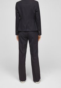 Black tailored suit jacket and pants. Smooth fabric with a fitted design, long sleeves, and buttons at the cuff. Worn with white sneakers.