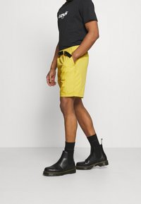 Žluté šortky s opaskem, bočními kapsami, v kombinaci s černými chelsea botami a černými ponožkami. Na vrchu je černé tričko s bílým potiskem.