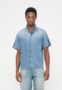 Πουκάμισο από τζιν με κοντά μανίκια σε ανοιχτό μπλε, με κουμπιά στο μπροστινό μέρος, γιακά και αντιθετικές σκουρότερες τζιν λεπτομέρειες στους ώμους και τα μανίκια.