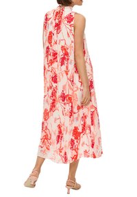 Robe fleurie en crème avec des motifs rouges et roses, sans manches, texture plissée et fermeture à nouer dans le dos. Portée avec des talons légers roses.