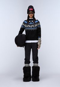 Czarny sweter z dzianiny z niebieskimi i białymi wzorami, czarne spodnie, szare akcenty skarpet, oraz czarne futrzane botki. Trzyma okrągłą czarną torbę.