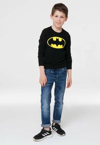 Schwarzes Langarmshirt mit gelbem Batman-Logo, kombiniert mit blauen Jeans und schwarzen Sneakers mit weißen Akzenten. Lässiges Freizeitoutfit für Jugendliche.