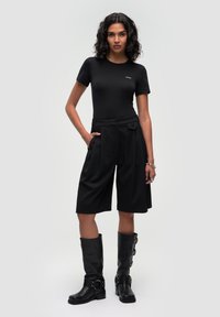 Černé přiléhavé tričko, střižené černé culoty s sklady a černé kožené vysoké boty s přezkami. Modelka má curly vlasy.
