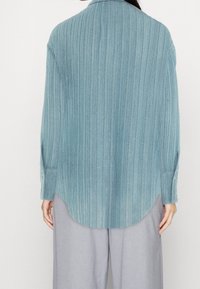 Jasnoniebieska, textured koszula z pionowymi paskami, z długimi rękawami i zaokrąglonym dołem. Wykonana z błyszczącego materiału.