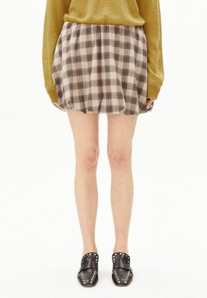 Personne portant un pull jaune, une jupe courte à carreaux beige et marron, et des chaussures slip-on noires cloutées, debout sur un fond blanc.