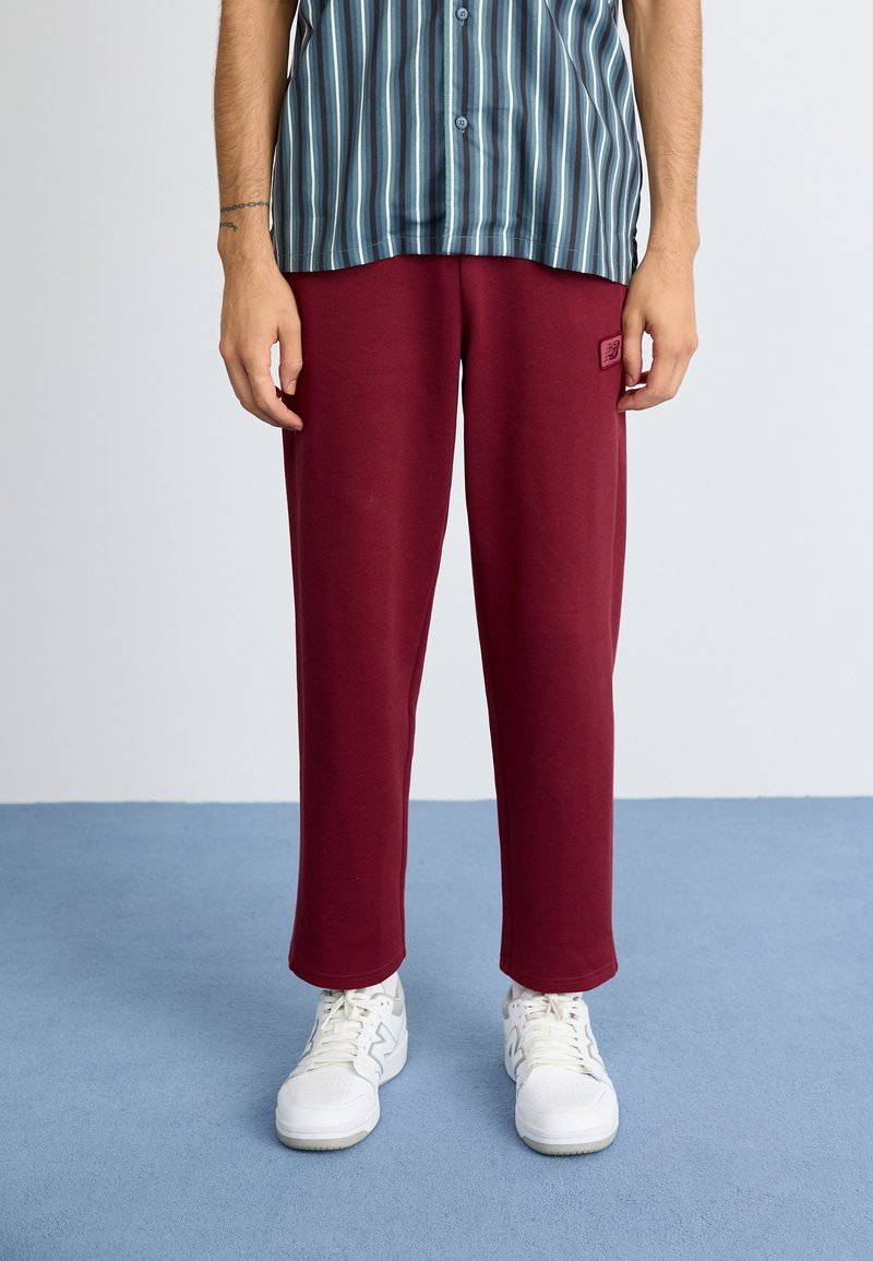 Pantaloni burgundy cu un croi lejer, realizați dintr-un material moale, având un mic patch cu logo pe coapsă și asortați cu teneși albi.