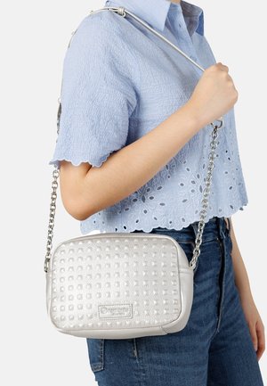 Frau trägt ein hellblaues, strukturiertes Kurzarmoberteil und Jeans, dazu eine kleine silberne, strukturierte Umhängetasche mit Kettenriemen.