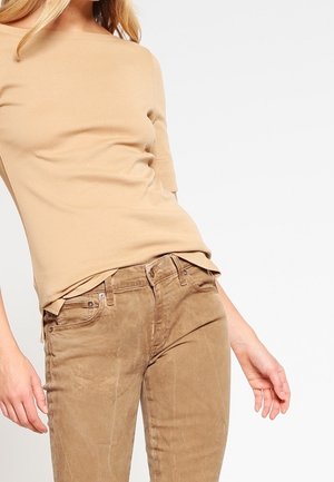 Kobieta ubrana w beżową bluzkę z krótkim rękawem i jasnobrązowe sztruksowe spodnie, stojąca z rękami swobodnie opuszczonymi wzdłuż ciała na jednolitym tle.