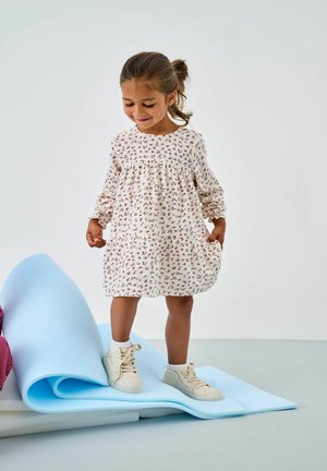 Beżowa sukienka w kwiatowy wzór z długimi rękawami i marszczonym dekoltem; dopasowana z białymi trampkami, stojąca na niebieskiej piance.