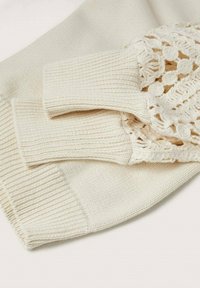 Pull en maille couleur crème avec poignets côtelés et détail en dentelle crochetée sur une manche, posé à plat sur une surface claire.