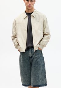 Lichtbeige bomberjack met een kraag, gecombineerd met blauwe denim shorts. Het jack heeft een ritssluiting aan de voorkant en zijzakken.