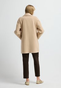Beige oversized jas met een grote kraag en textuur mouwen, gedragen over bruine broeken en gouden instappers, vanuit de achterkant bekeken.